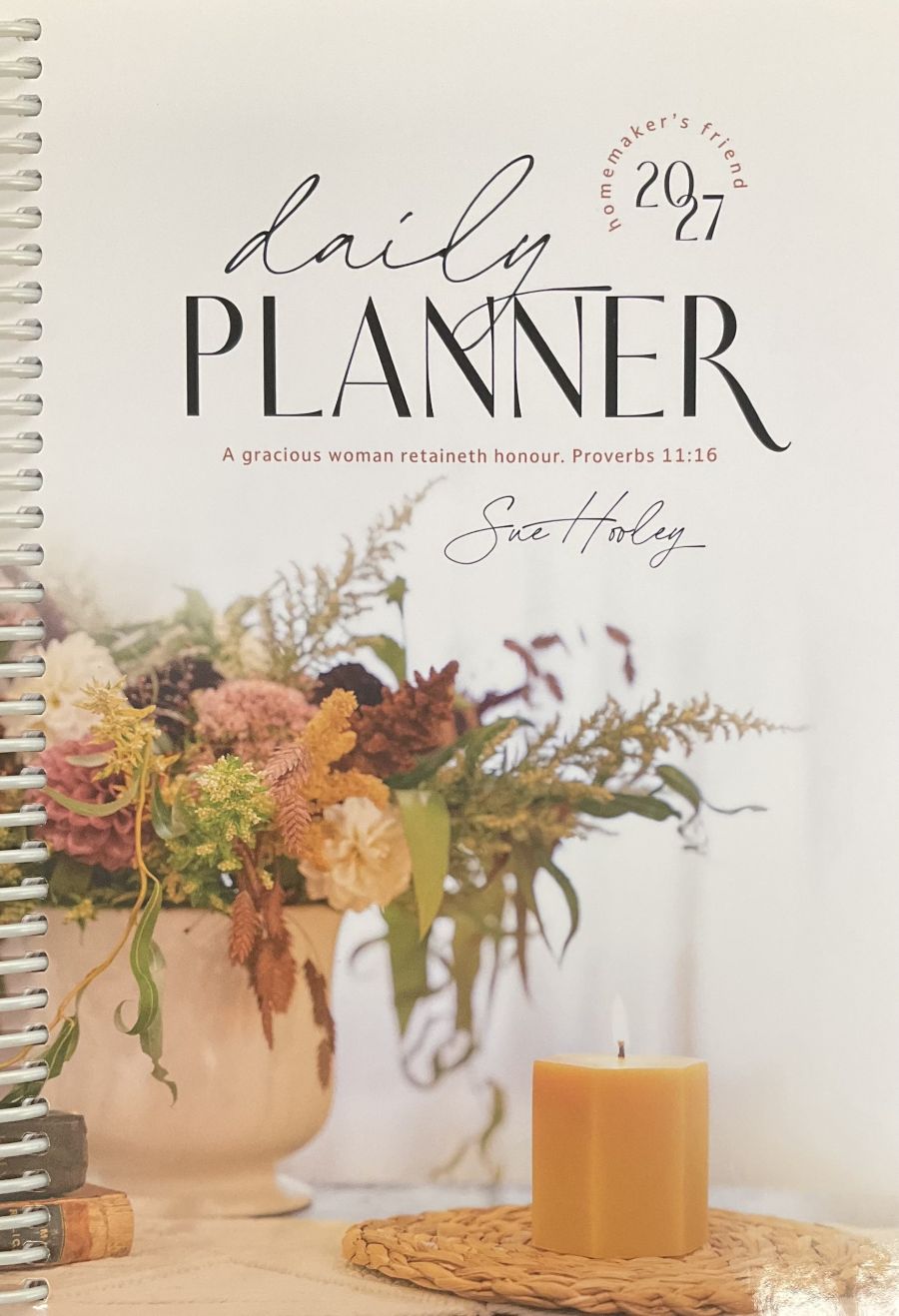 A spiral-bound daily planner cover shown flat, featuring the title 'daily PLANNER' and '2027' with a quote from Proverbs 11:16. The cover includes a floral arrangement and a candle in a warm, soft-toned design.