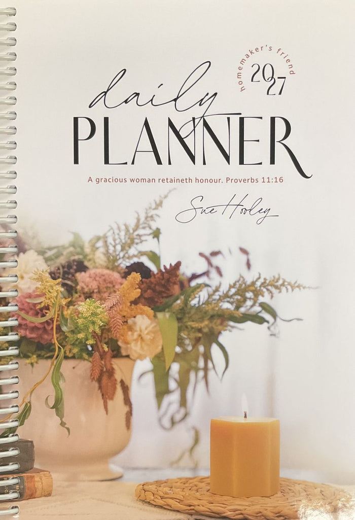 A spiral-bound daily planner cover shown flat, featuring the title 'daily PLANNER' and '2027' with a quote from Proverbs 11:16. The cover includes a floral arrangement and a candle in a warm, soft-toned design.